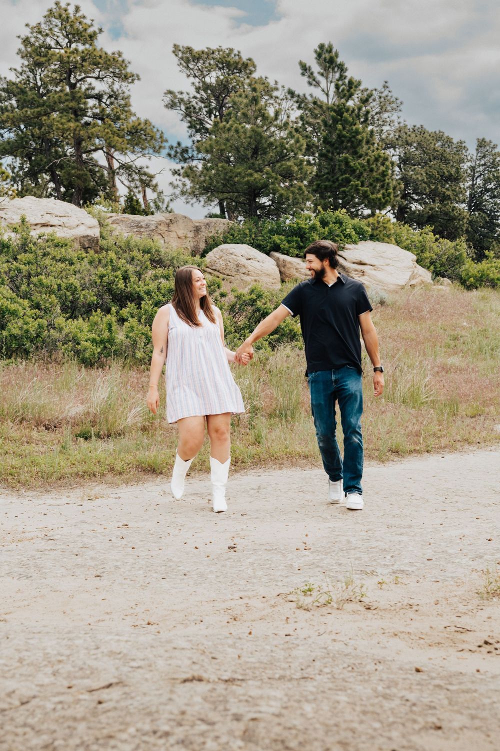 A couple walks away from the camera on a gravel path surrounded by green trees and grass on a cloudy day.