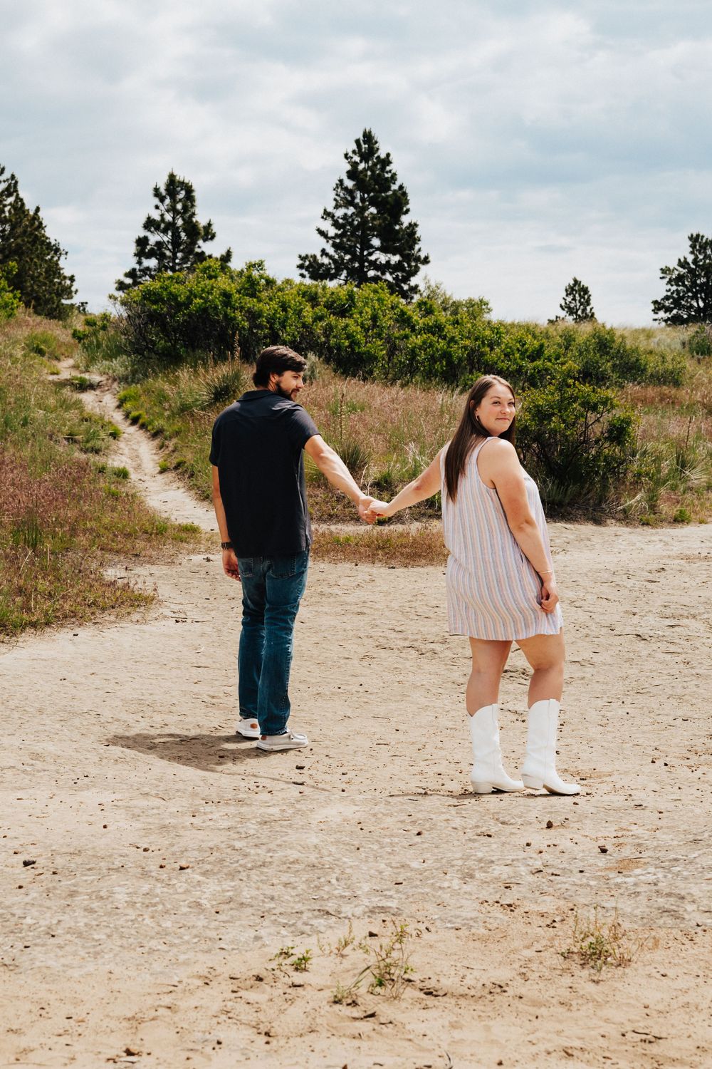 A couple walks hand in hand along a sandy nature trail surrounded by trees and brush on a cloudy day.