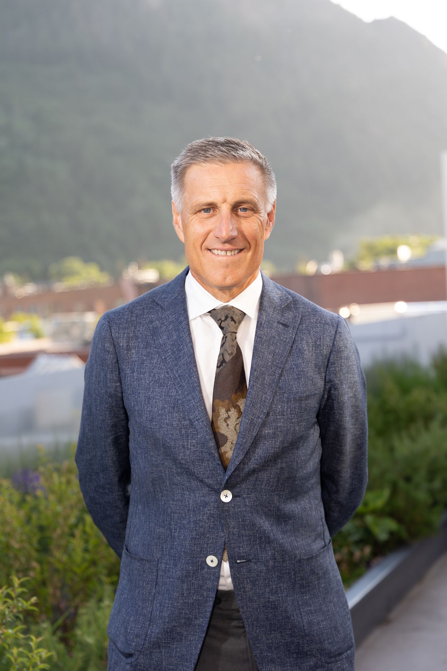 Professional portrait of someone wearing a navy blue suit and patterned tie against an outdoor background.