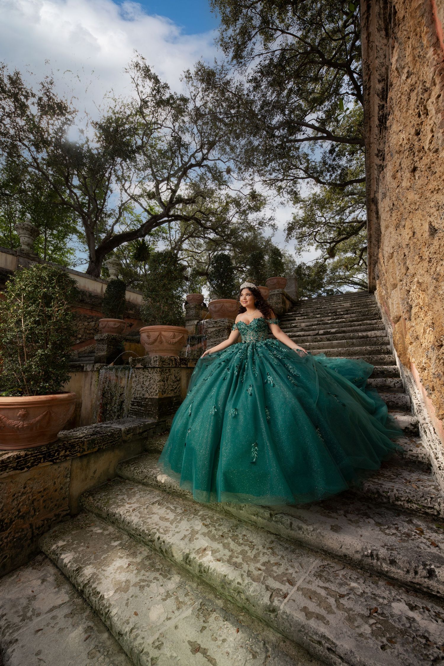 A person in an emerald green ball gown poses gracefully on stone steps in an outdoor garden setting.