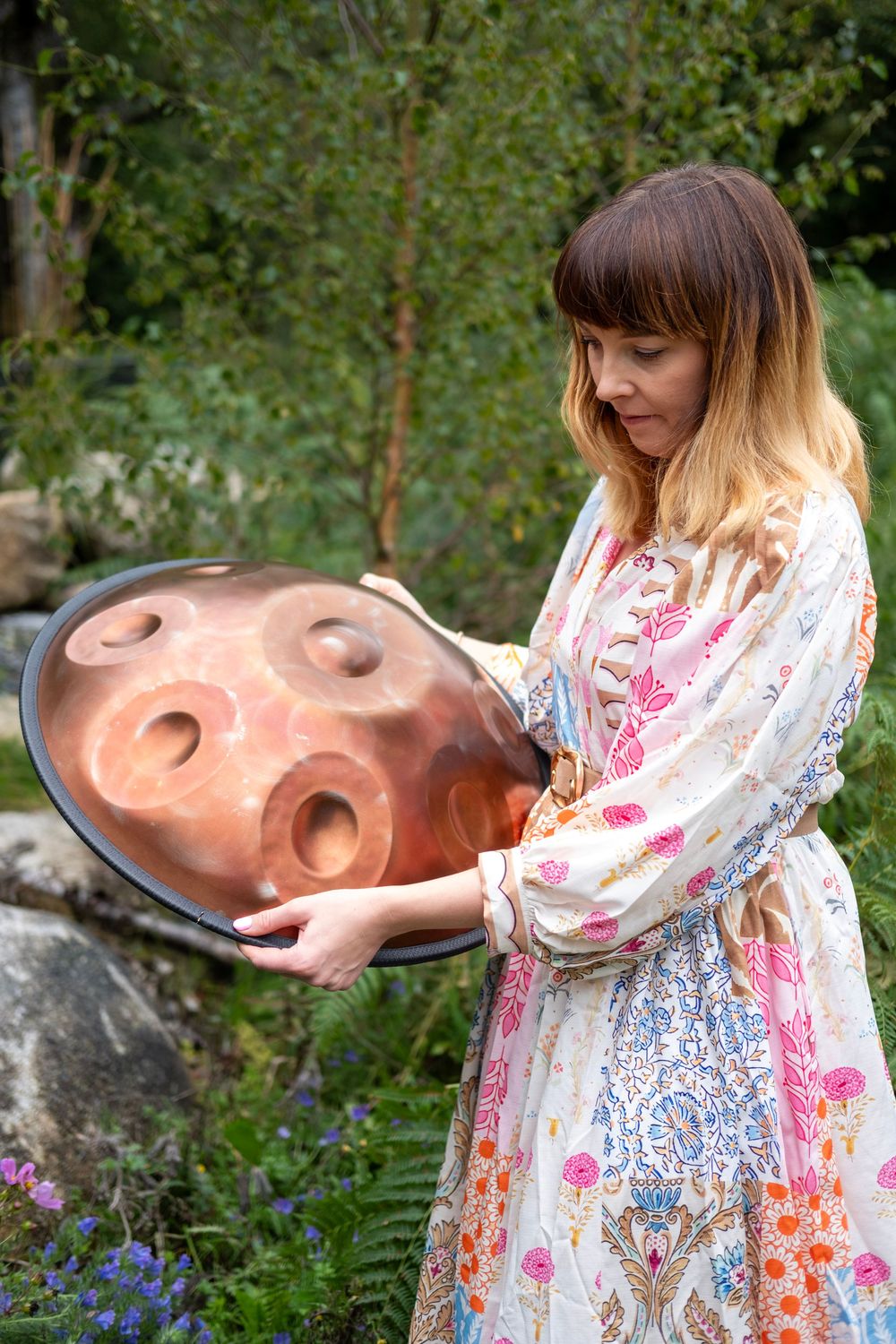A person in a floral dress interacts with a large sculptural face in an outdoor garden setting.