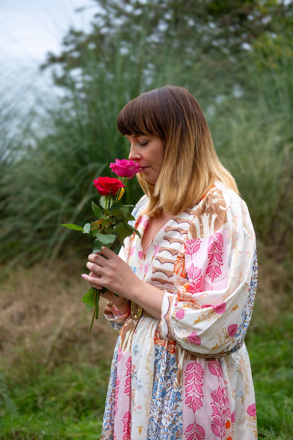 A person in a floral dress smells a red rose in a garden setting.