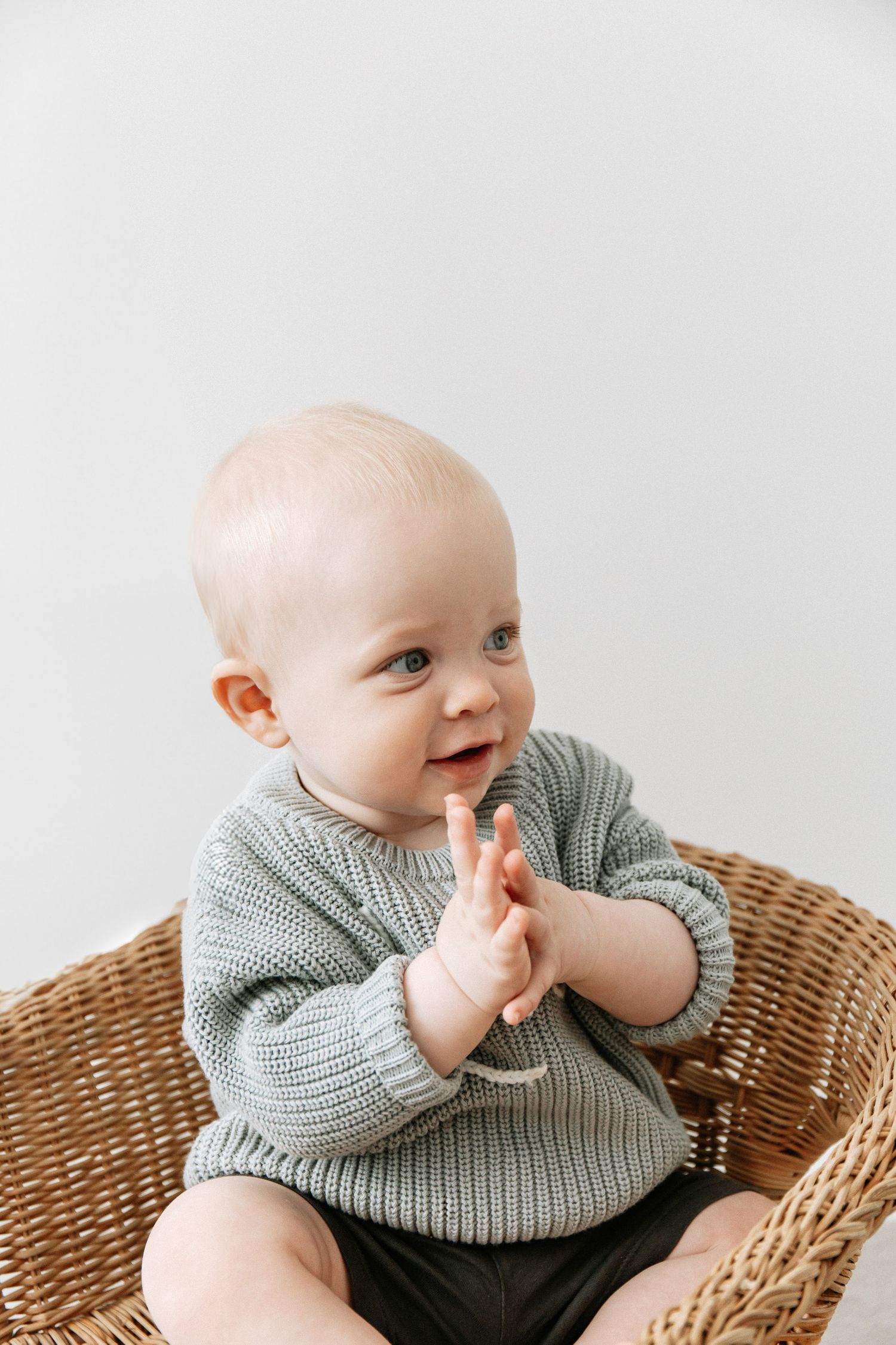 A baby wearing a blue knit jumper sits in a wicker chair and smiles while clapping with their hands, during birthday shoot in Nottingham