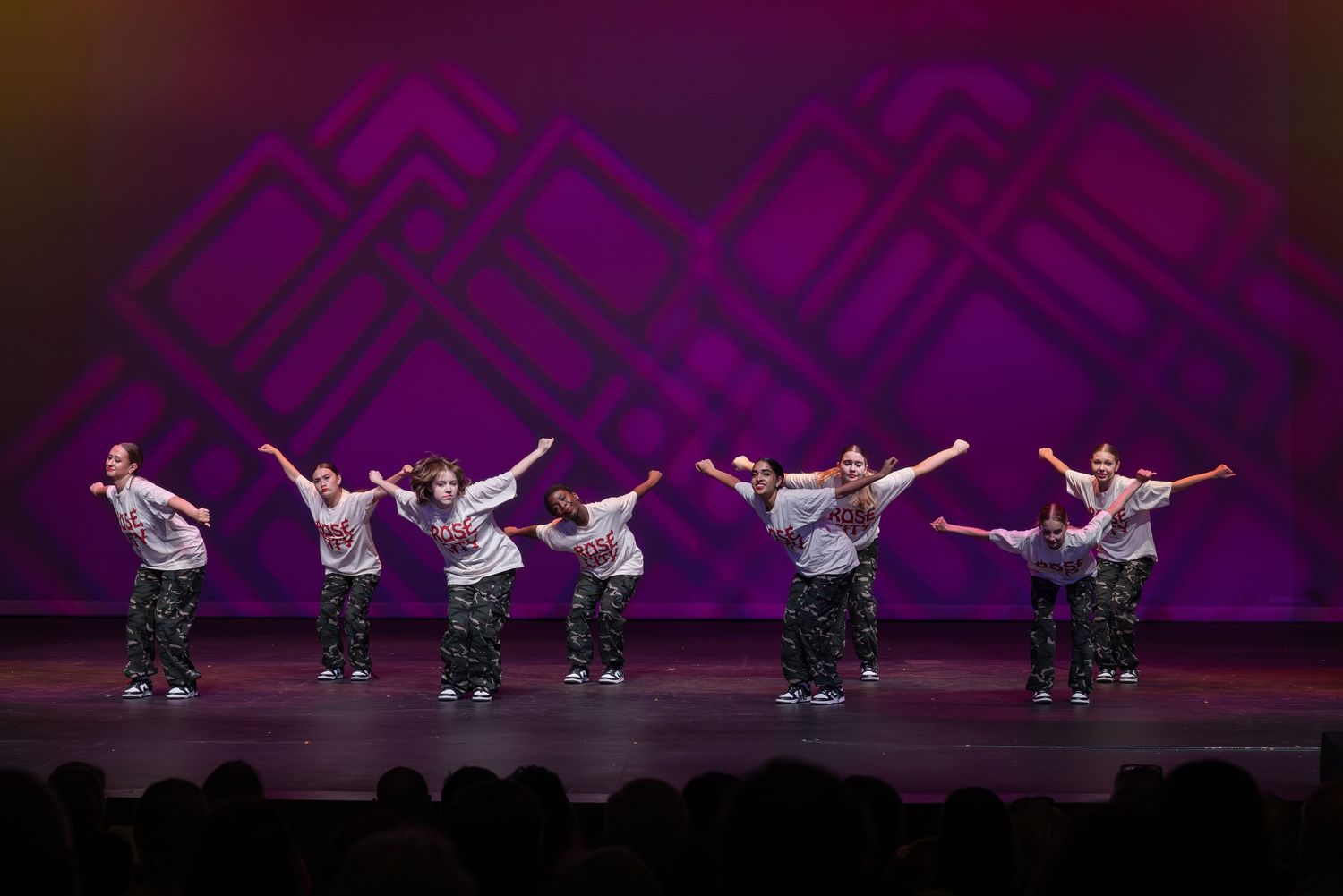Dancers perform a synchronized leap sequence on stage with dramatic purple lighting and dark silhouettes.