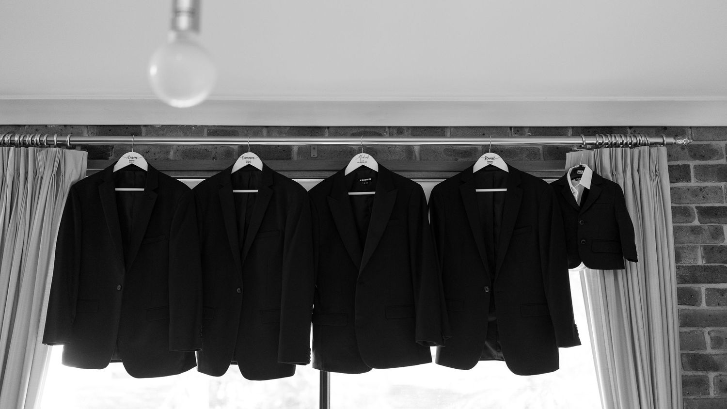 Five black formal suit jackets hanging in a row on white hangers against a brick wall.