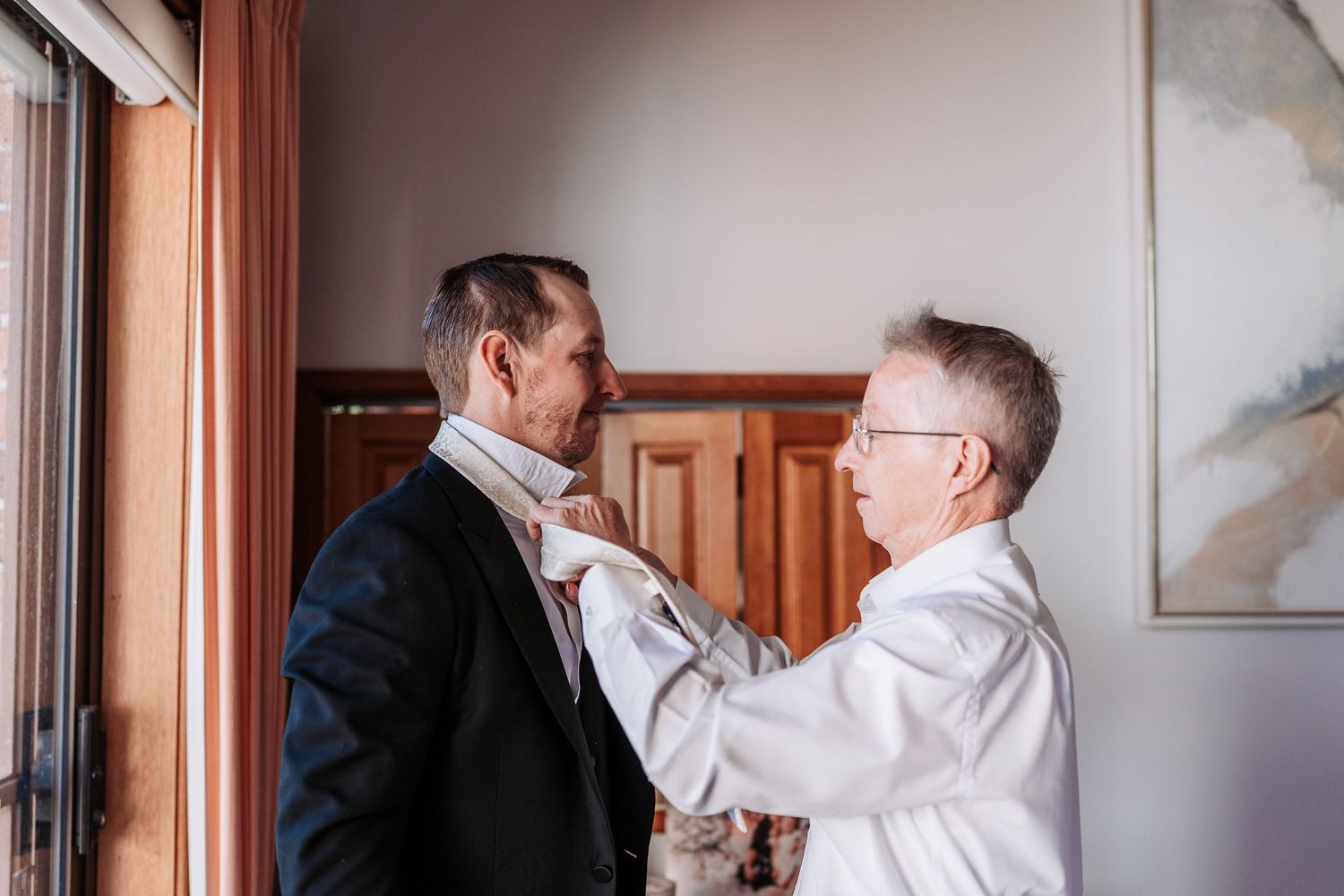 Two people in formal attire help adjust a tie and prepare for a special event in an elegant room.