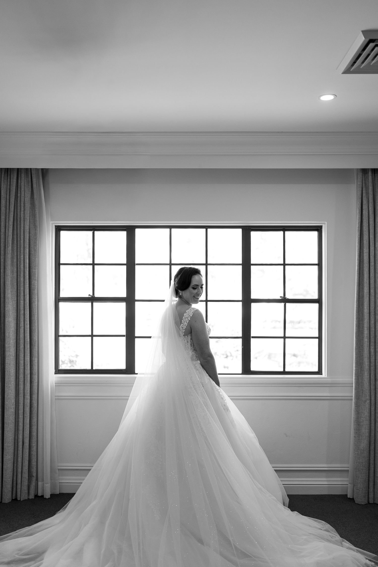 Elegant bridal portrait in black and white showing a wedding dress with flowing train against large windows.