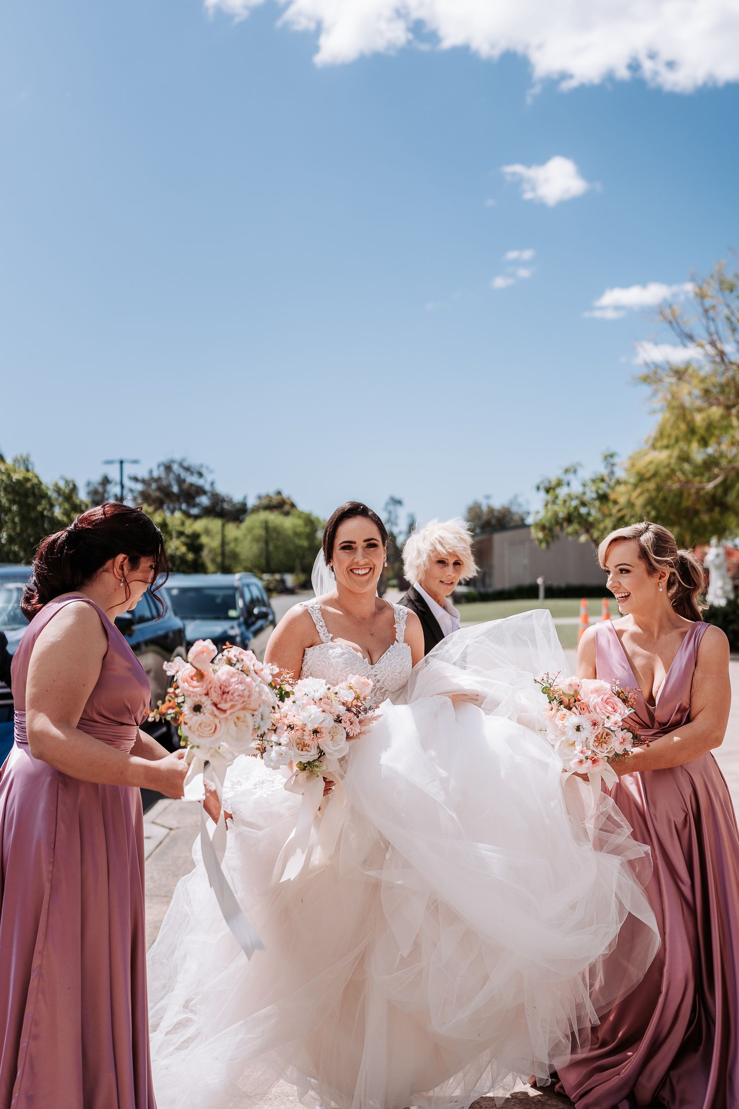 Bridesmaids in dusty rose dresses help adjust a flowing white wedding gown outdoors on a sunny day.