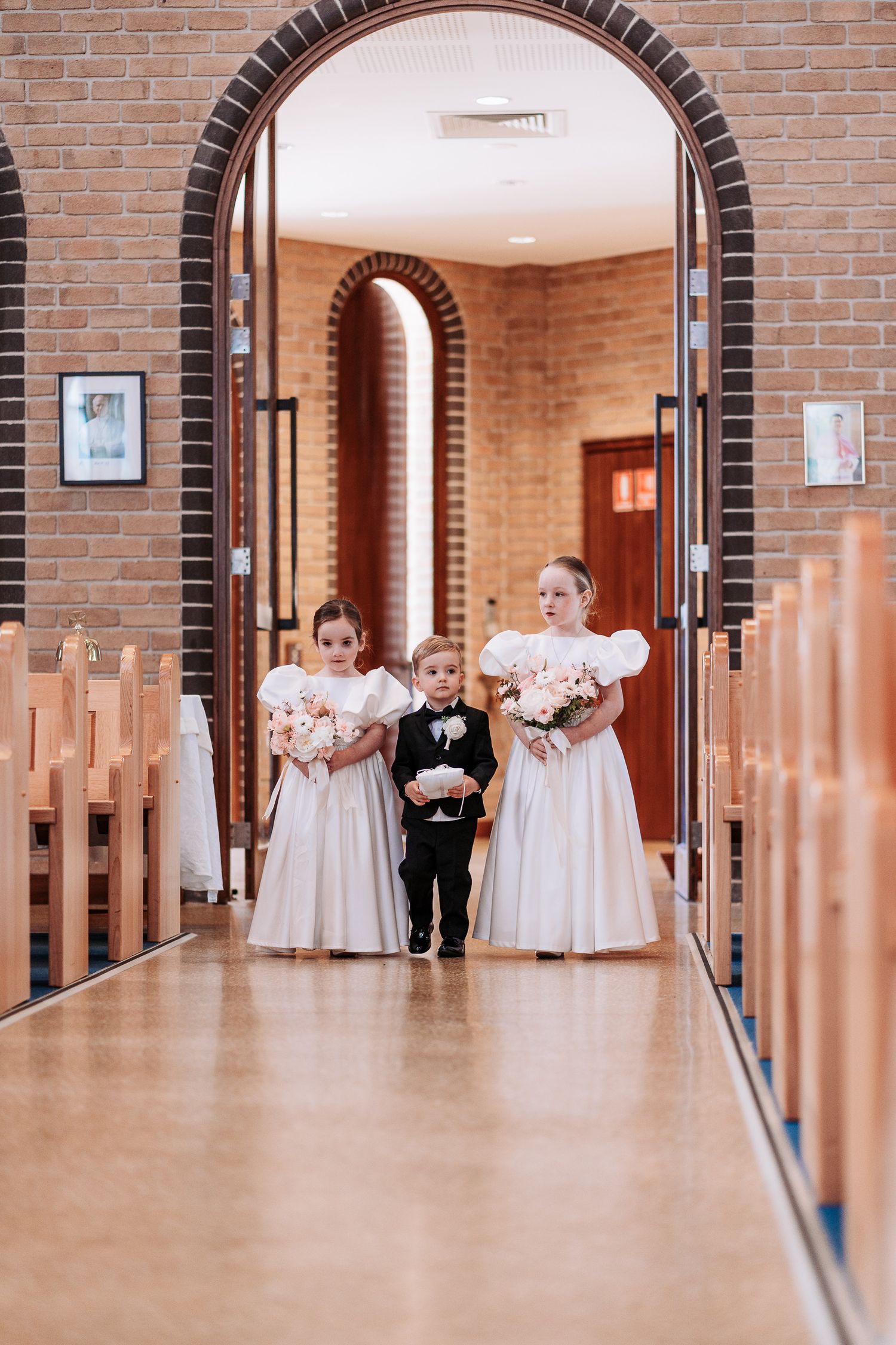 Wedding party members walk down a church hallway with brick walls and arched doorways.