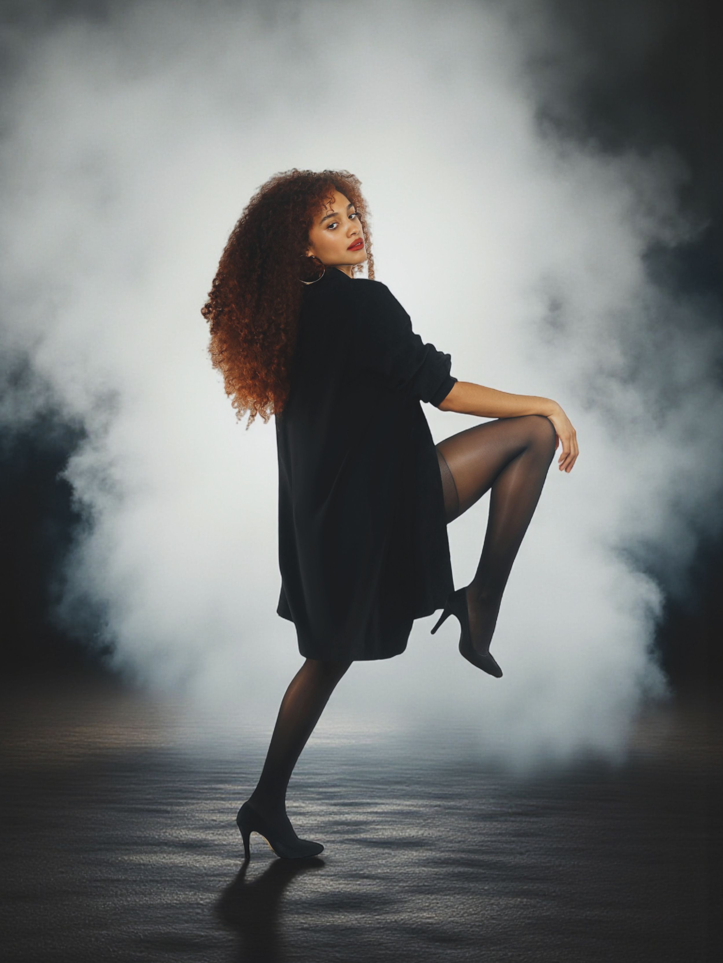 A dancer in a black dress performs an elegant leap through atmospheric fog on a dark stage.