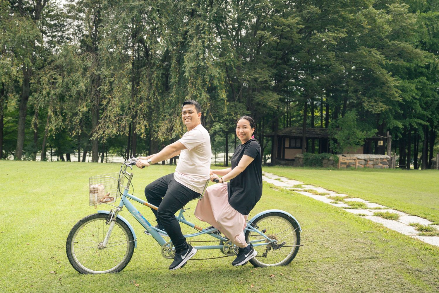 Two people ride together on a light blue tandem bicycle across a green lawn with trees in the background.