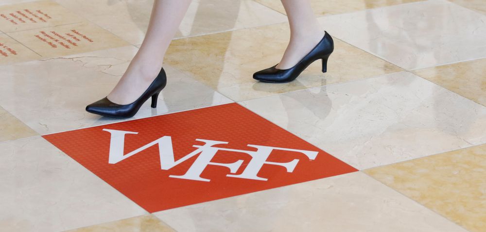 Two pairs of black high heels walking across a red WFF logo on a tiled floor.