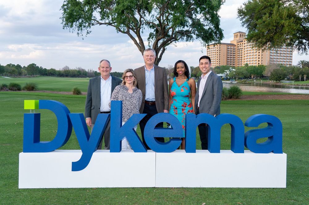 Outdoor corporate group photo with large trees and buildings in the background.