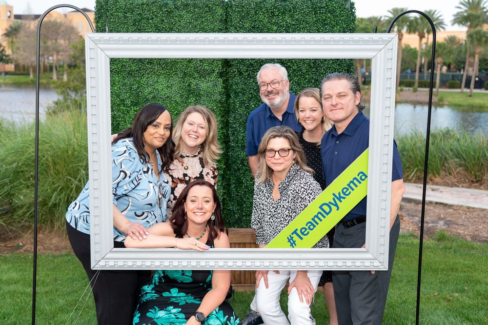A group poses together outdoors with a white decorative photo frame prop during a sunny day celebration.