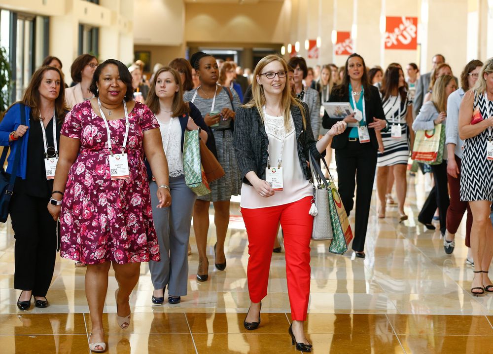 Large group of people walking through a bright indoor hallway with tiled floors.