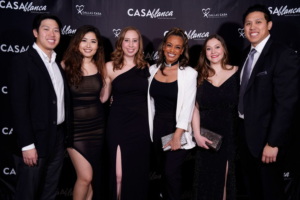 Group photo of attendees in formal black attire against branded step-and-repeat backdrop.