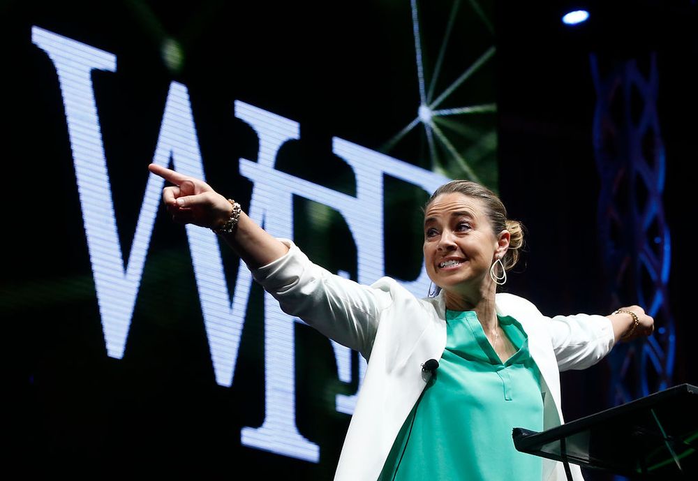 A speaker in a white blazer and turquoise top gestures enthusiastically on stage with illuminated text behind them.