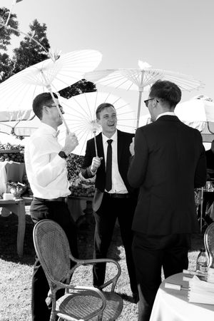 People holding white parasols gather outdoors at a formal event in black and white attire.