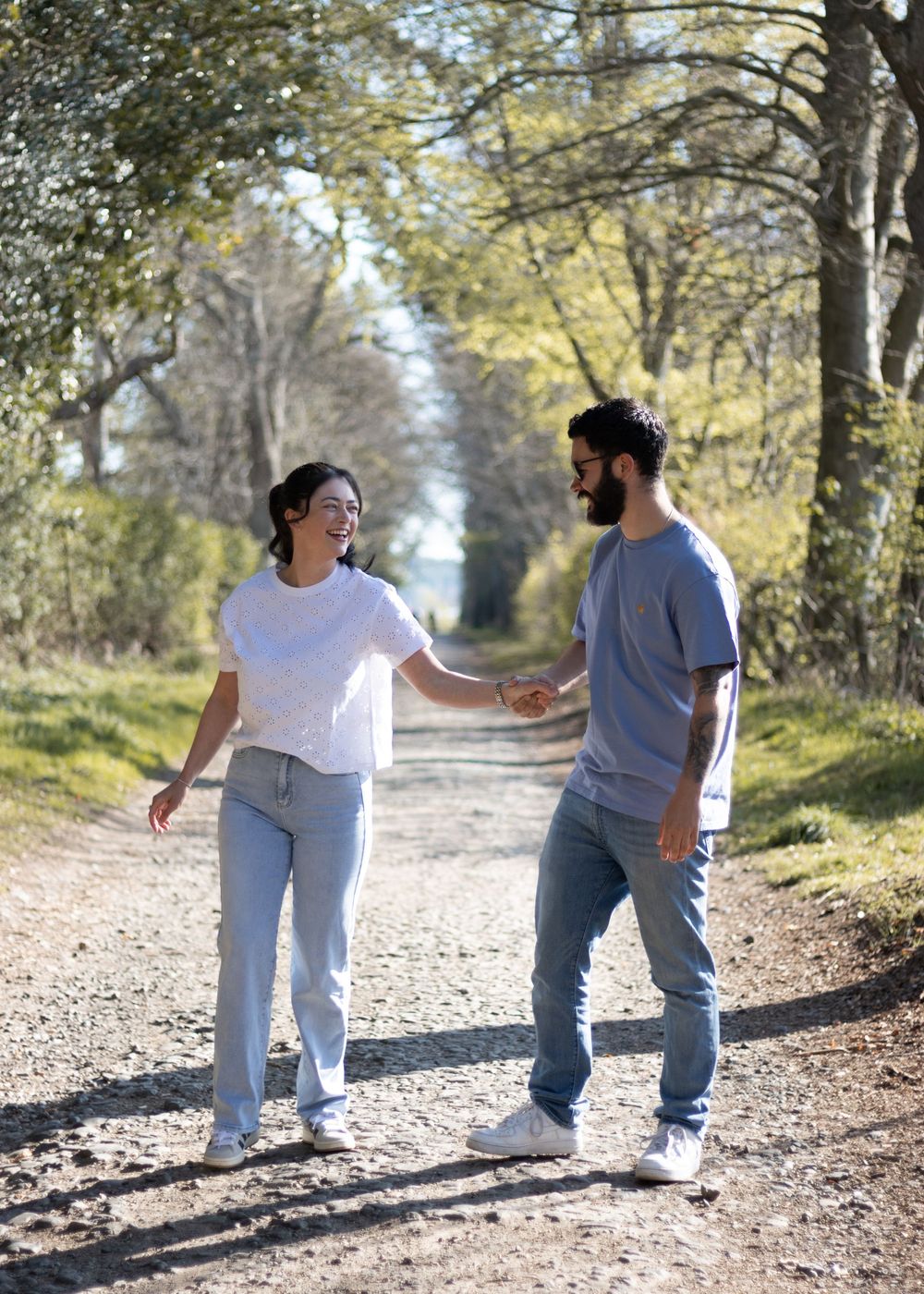 A couple walks together on a tree-lined dirt path during a sunny autumn day.