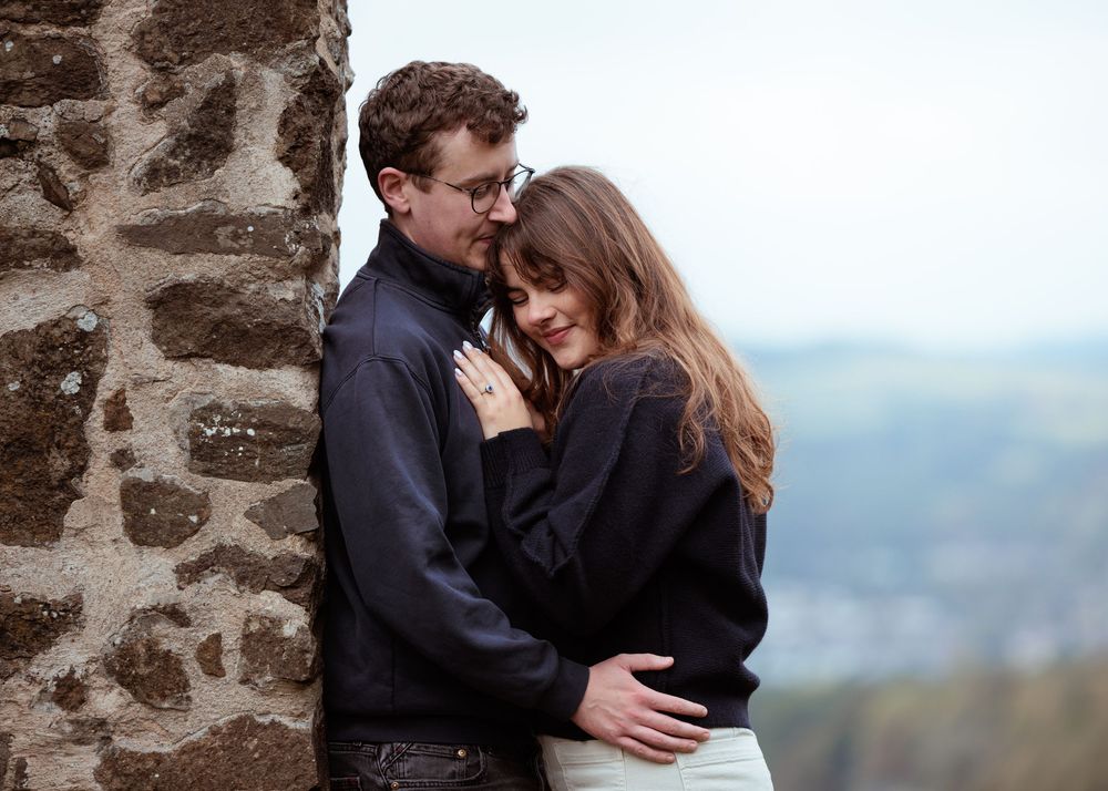 A romantic couple embraces against a stone wall overlooking a misty mountain landscape.