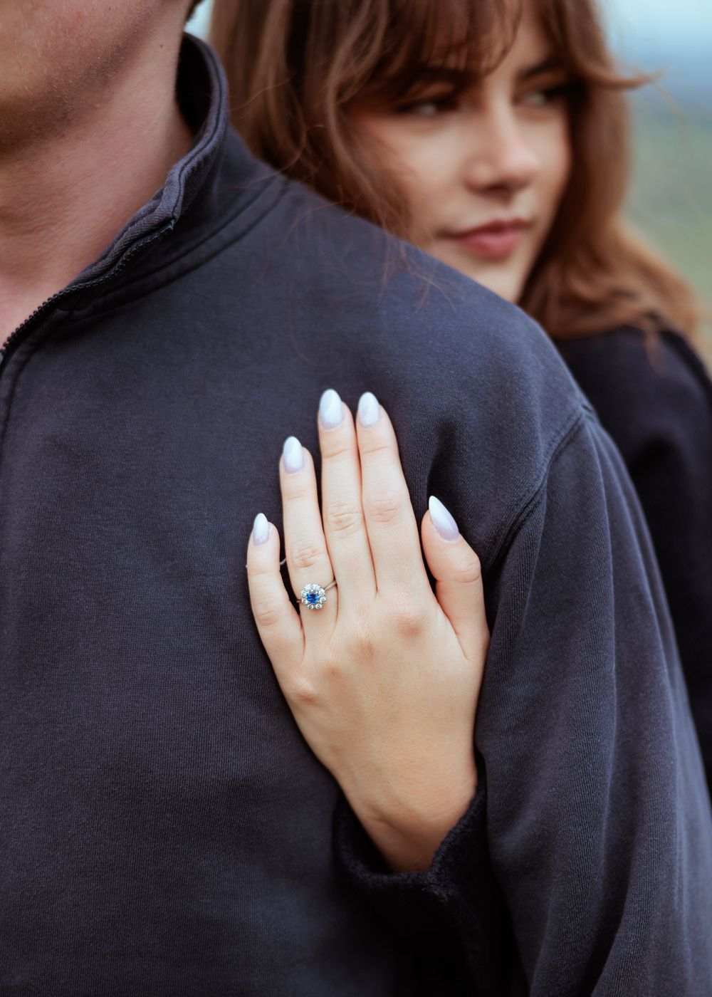 Close-up view of a diamond engagement ring on someone's hand against a dark background.
