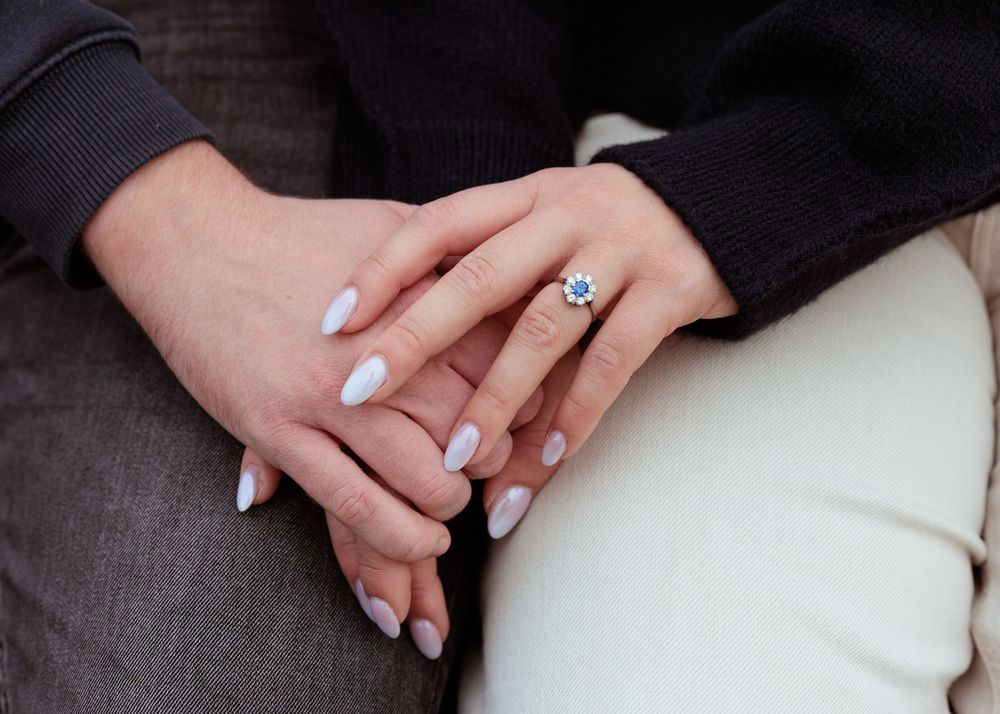 Close-up of hands with engagement ring resting on a grey couch showing wedding band and manicured nails.