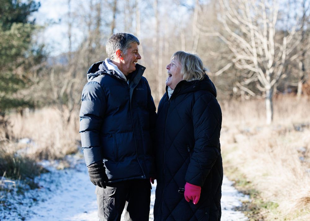 Two people in winter coats walking together through a snowy landscape with bare trees in the background.