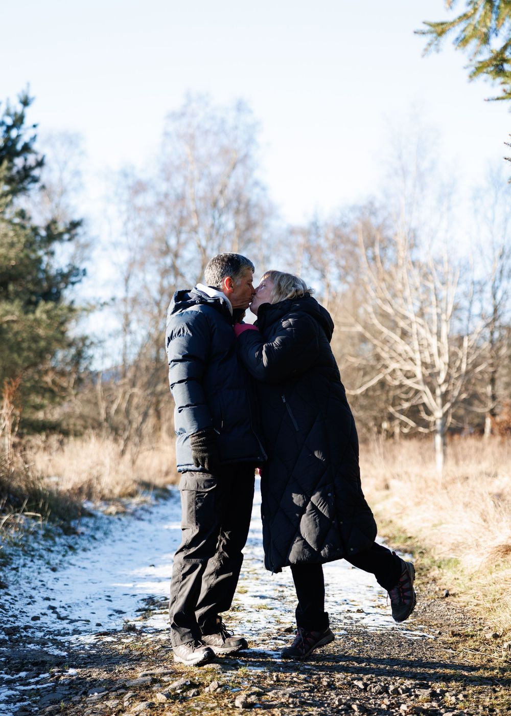 A couple in winter coats sharing a romantic moment on a snowy forest path with bare trees in the background.