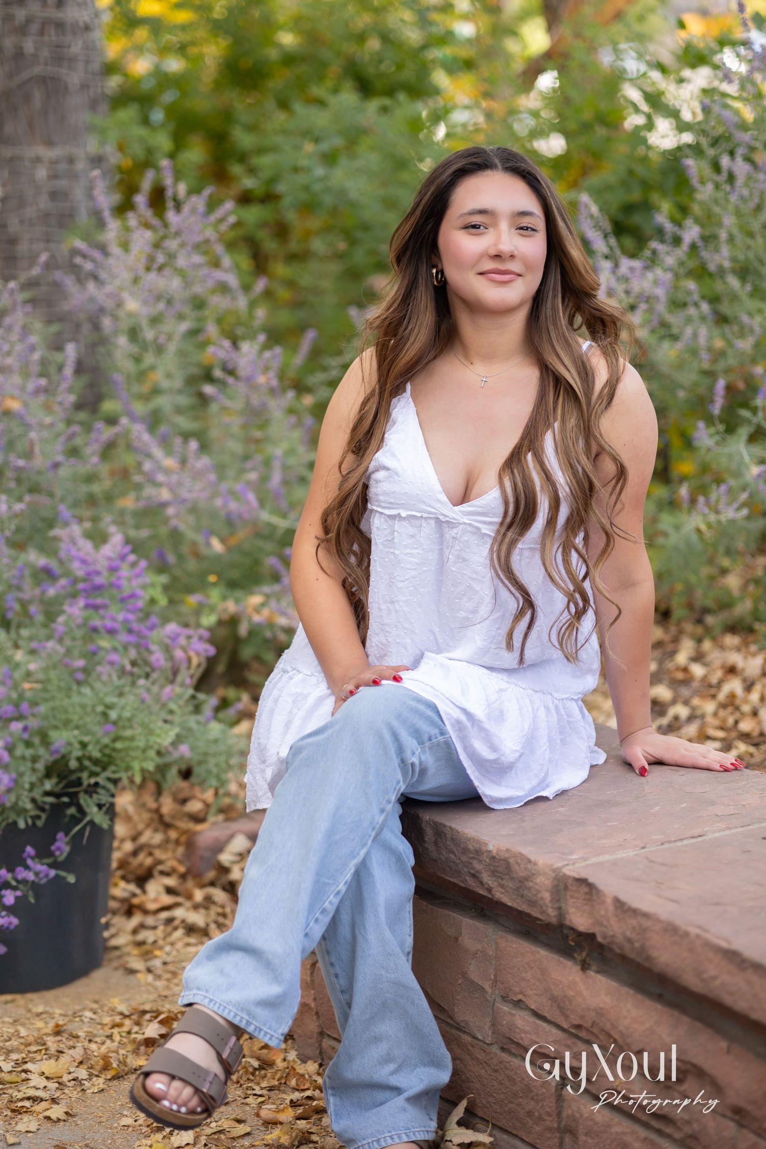 Model in white dress and jeans sits casually on stone wall surrounded by garden flowers.