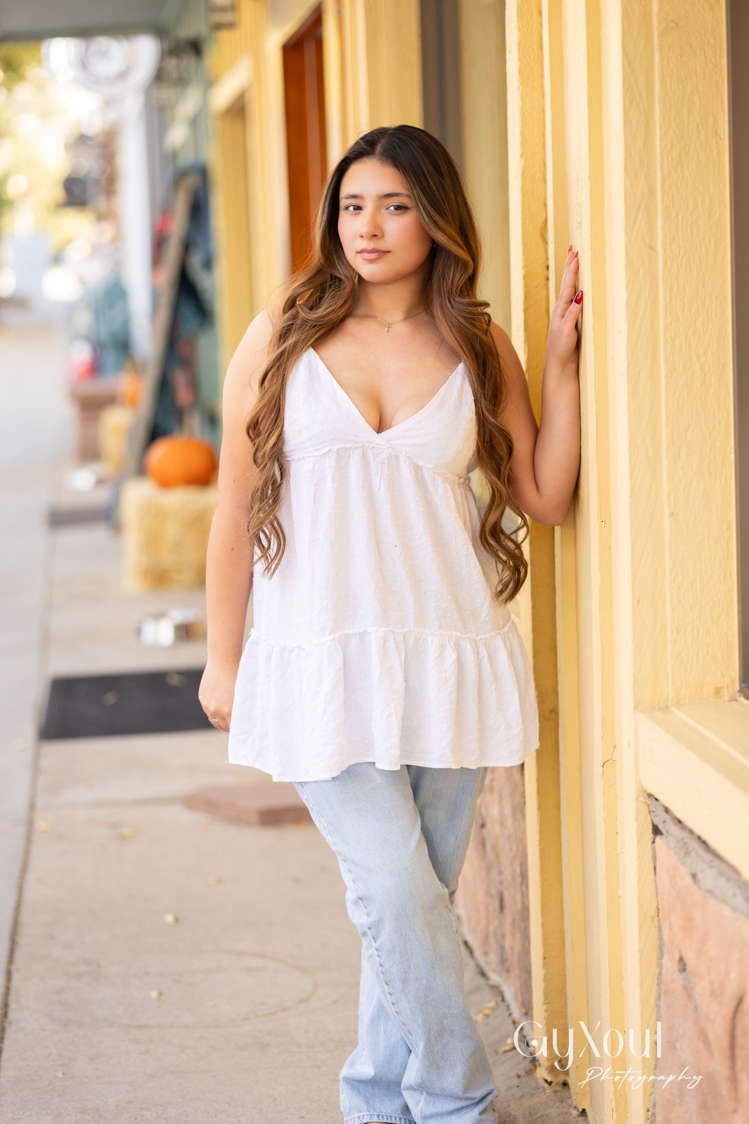 Model in white flowing top and jeans poses against yellow wall with pumpkin decorations.