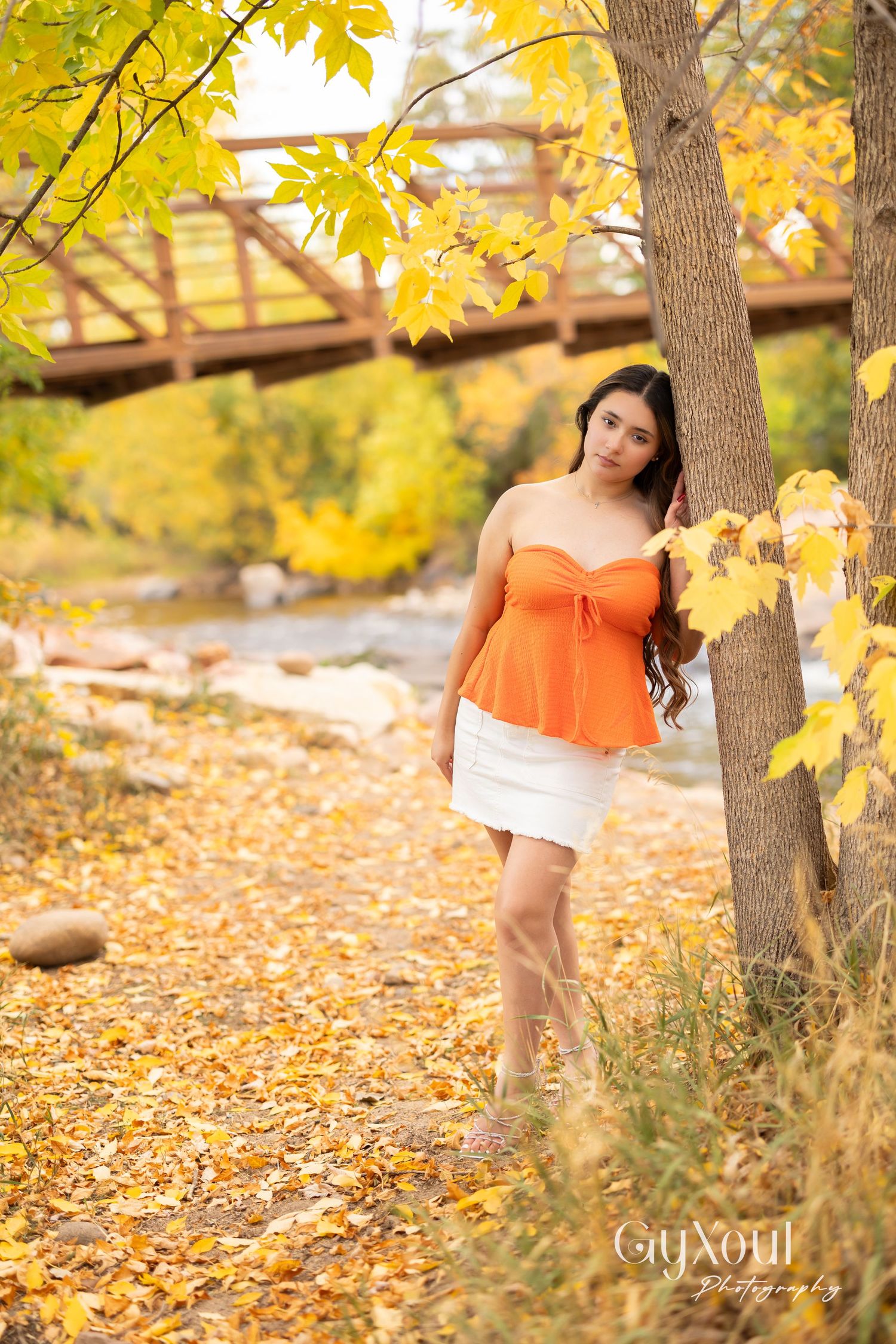 Figure in orange top and white skirt poses under yellow autumn leaves by wooden bridge.