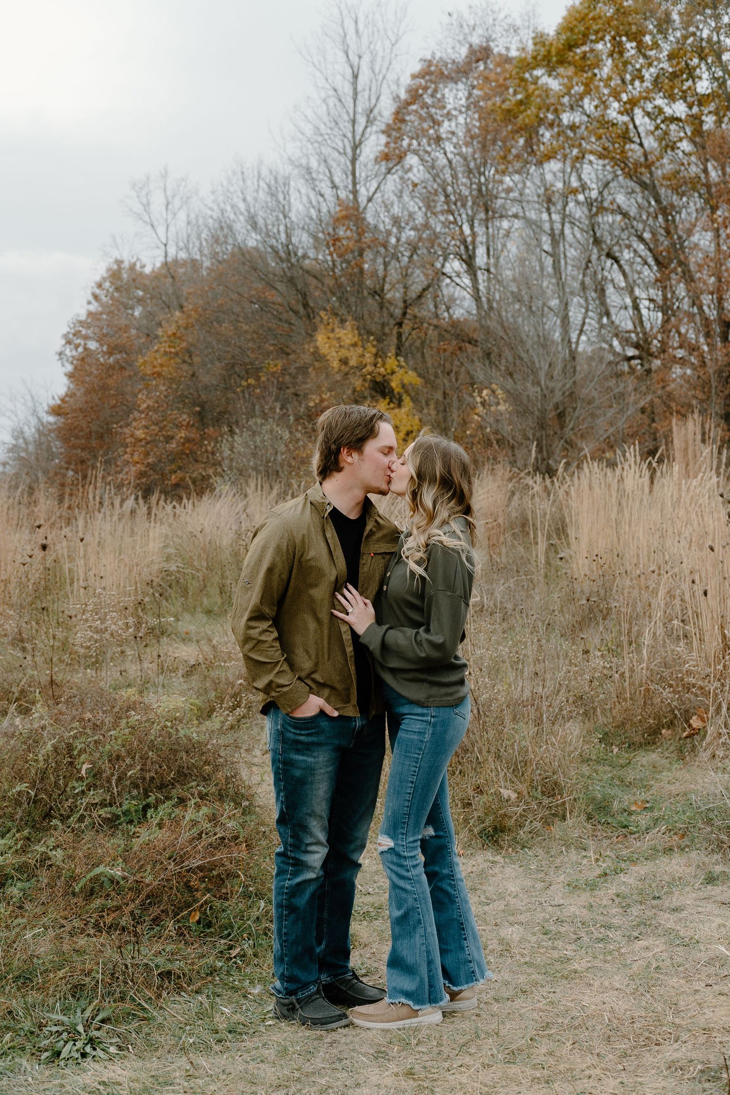 A couple embraces on a rustic dirt path surrounded by autumn foliage and tall dried grasses.
