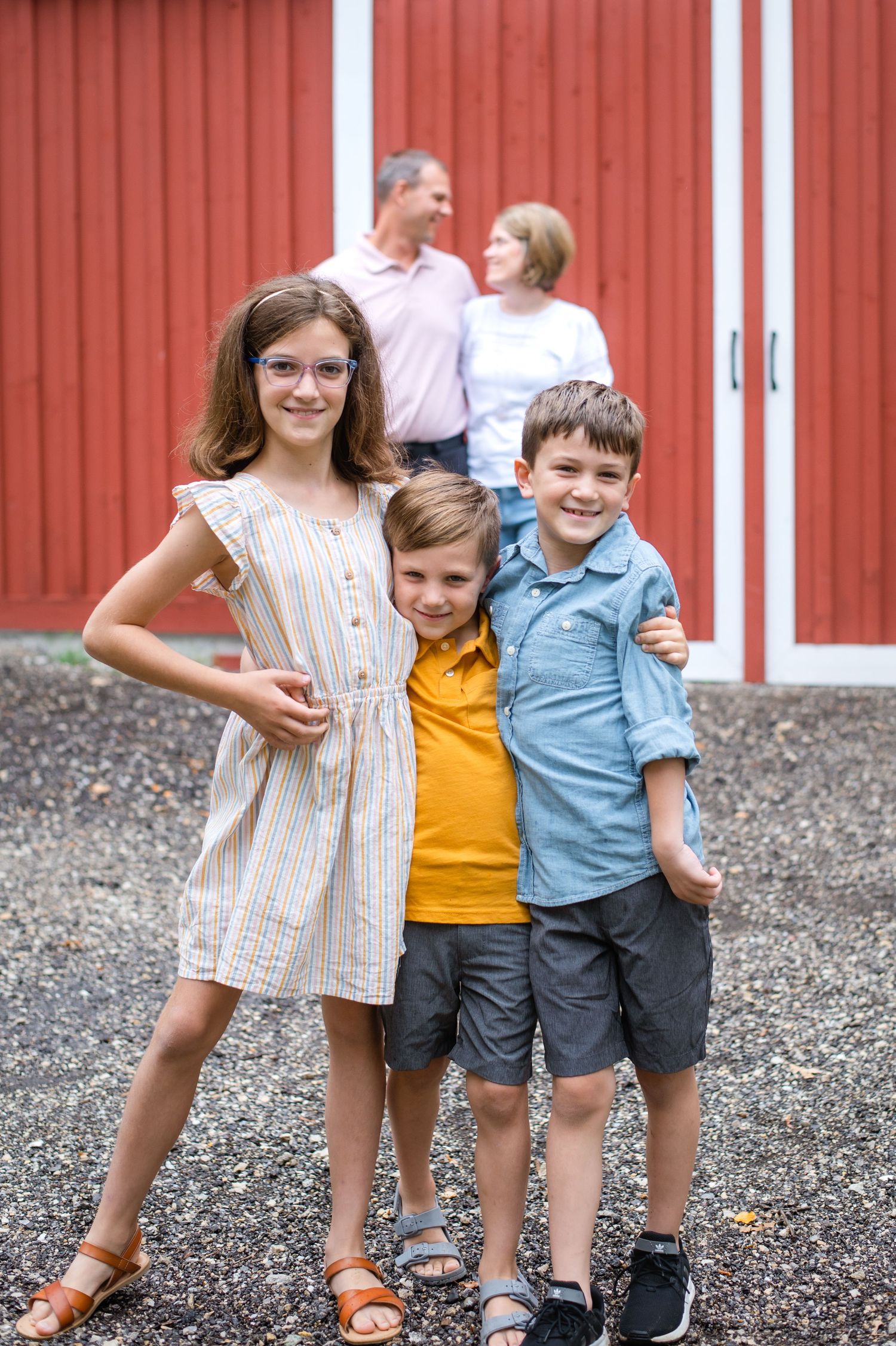 Children stand together laughing and playing in front of a red barn wall on a gravel surface.