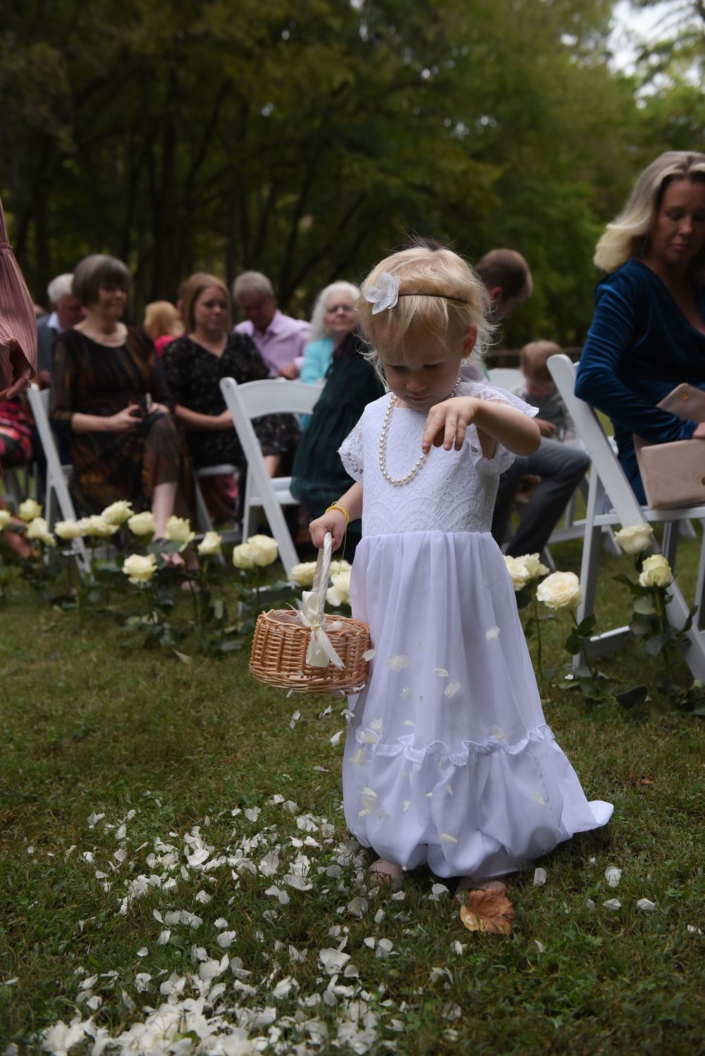 A small flower girl in a white dress scatters petals down the aisle at an outdoor wedding ceremony.