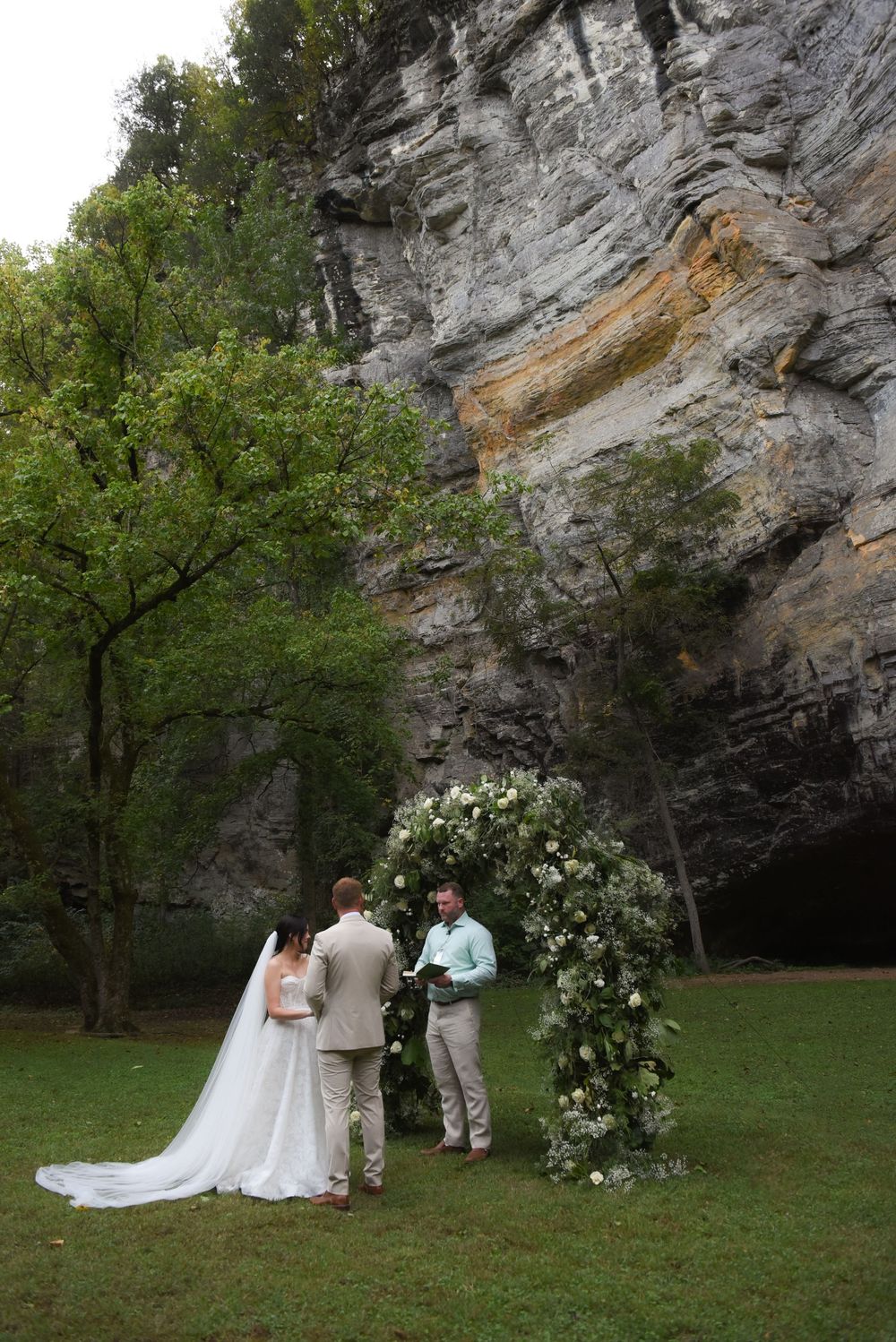 An intimate outdoor wedding ceremony takes place against a dramatic rock wall backdrop with white floral arrangements.