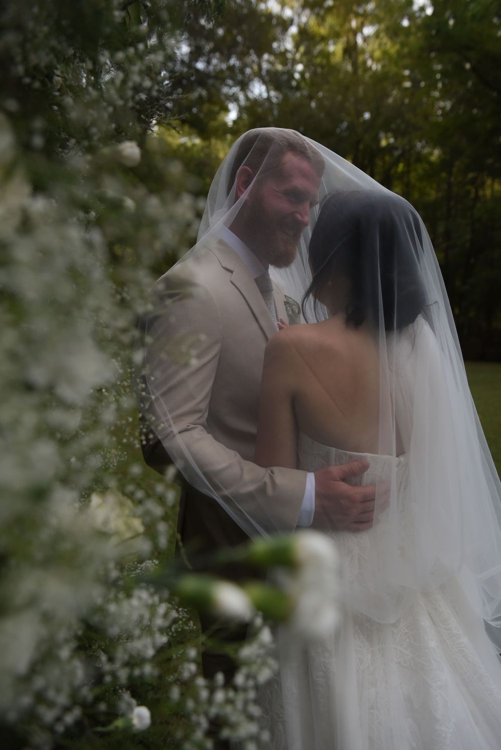 A romantic wedding couple shares an intimate moment outdoors framed by soft white flowers and greenery.