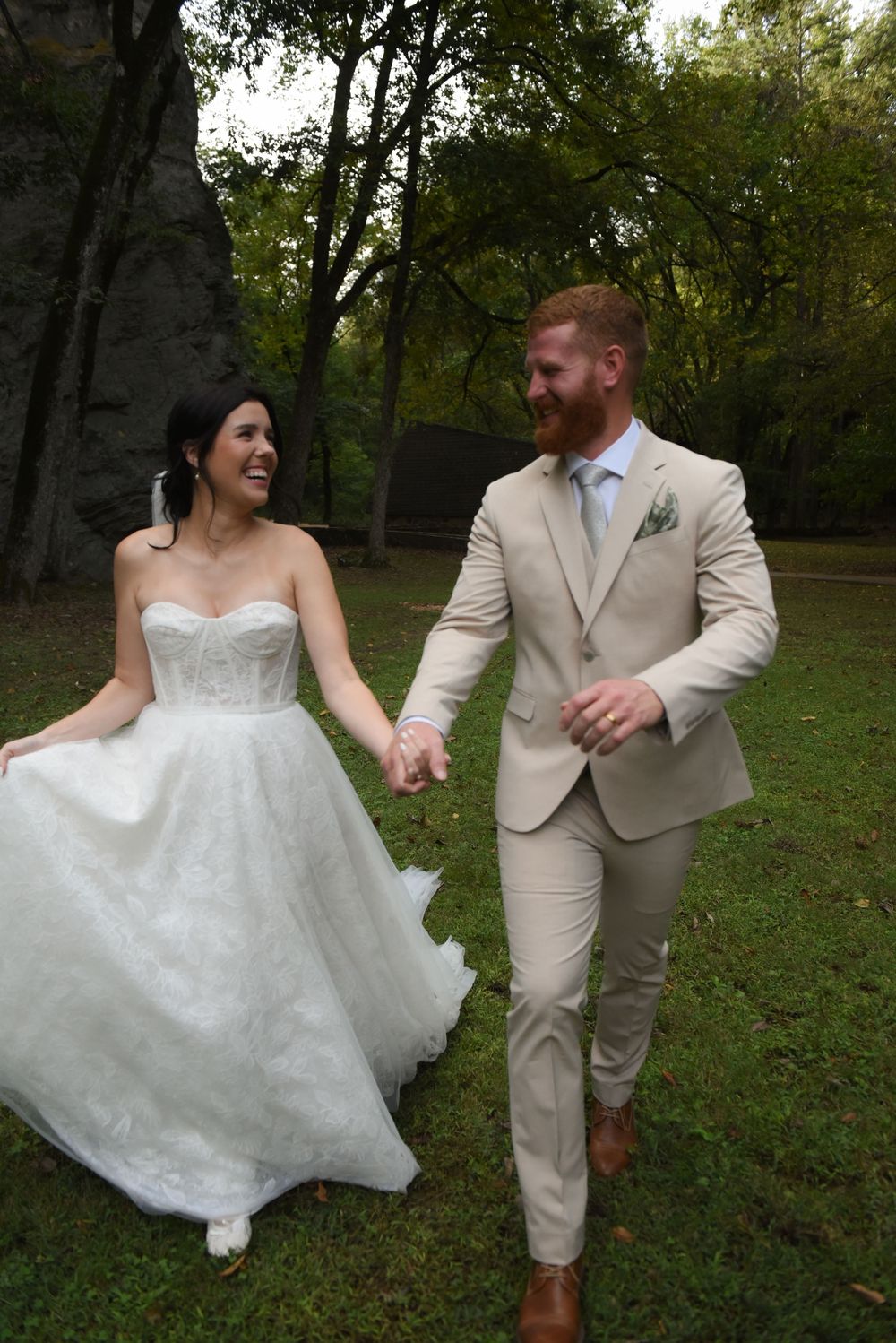 A bride and groom in a white dress and tan suit dance playfully in a wooded outdoor setting.
