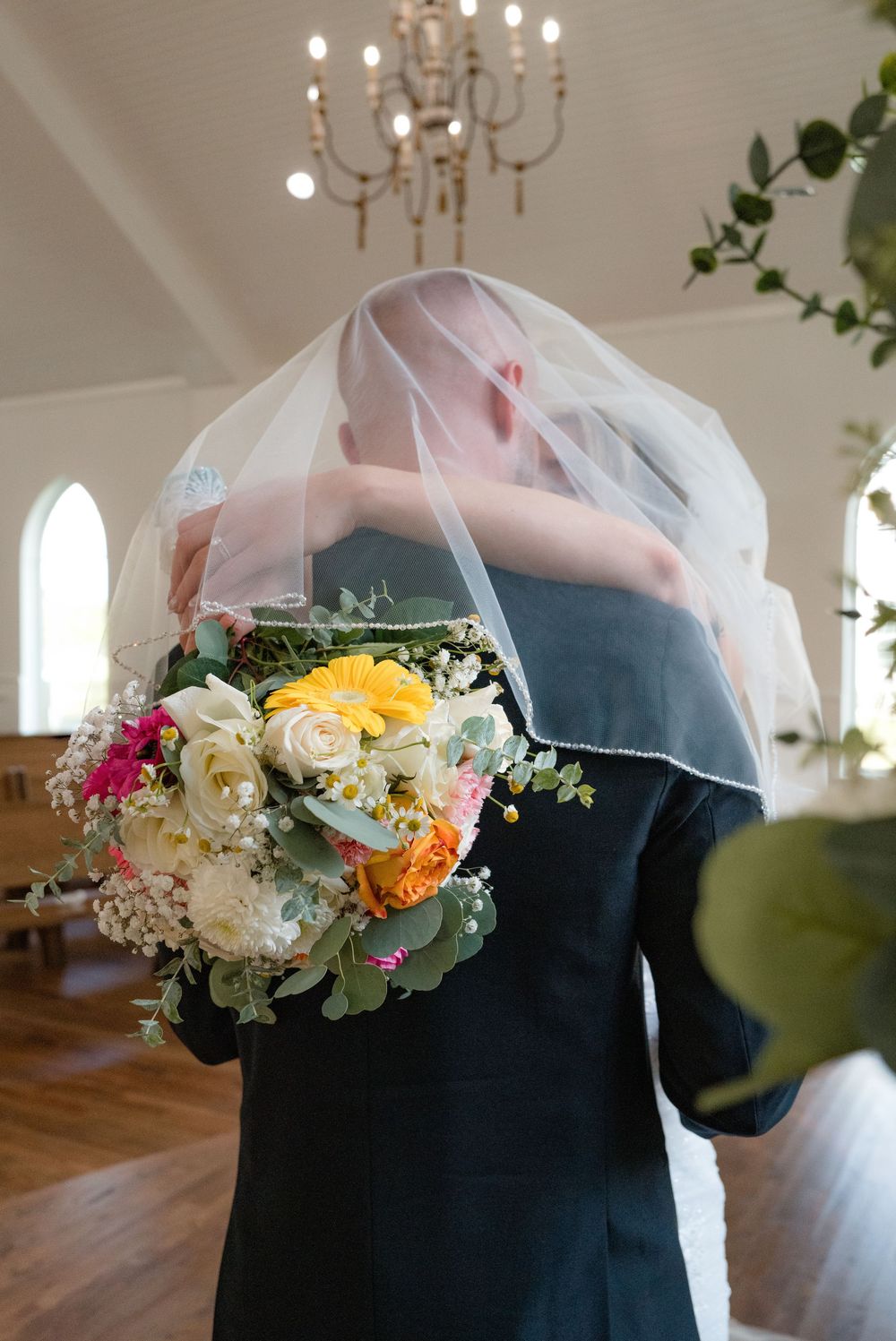 Floral bouquet being held against a dark dress in an elegant room with chandeliers and hardwood floors.