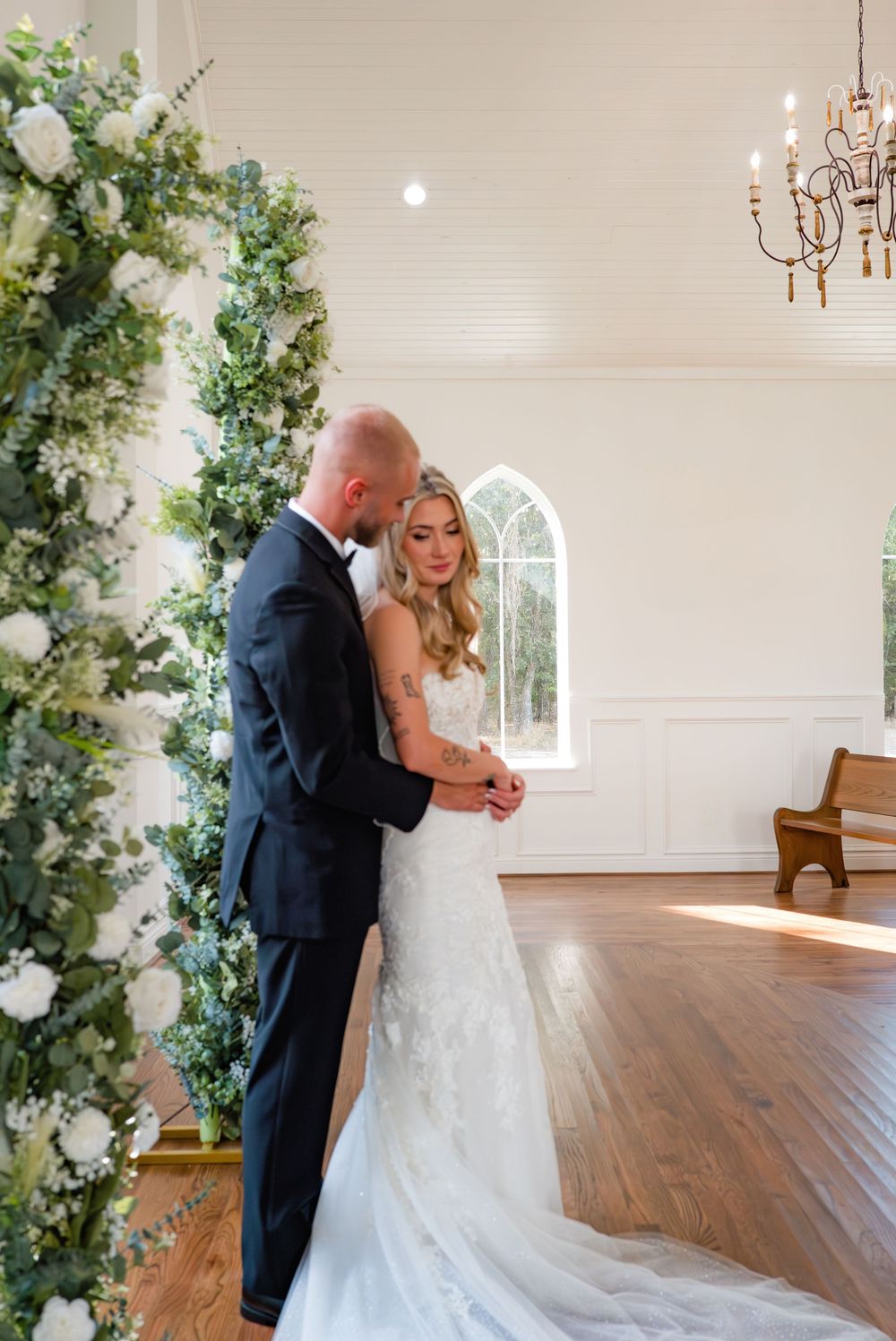 A romantic wedding couple shares a tender moment in an elegant white chapel with floral archways and hardwood floors.