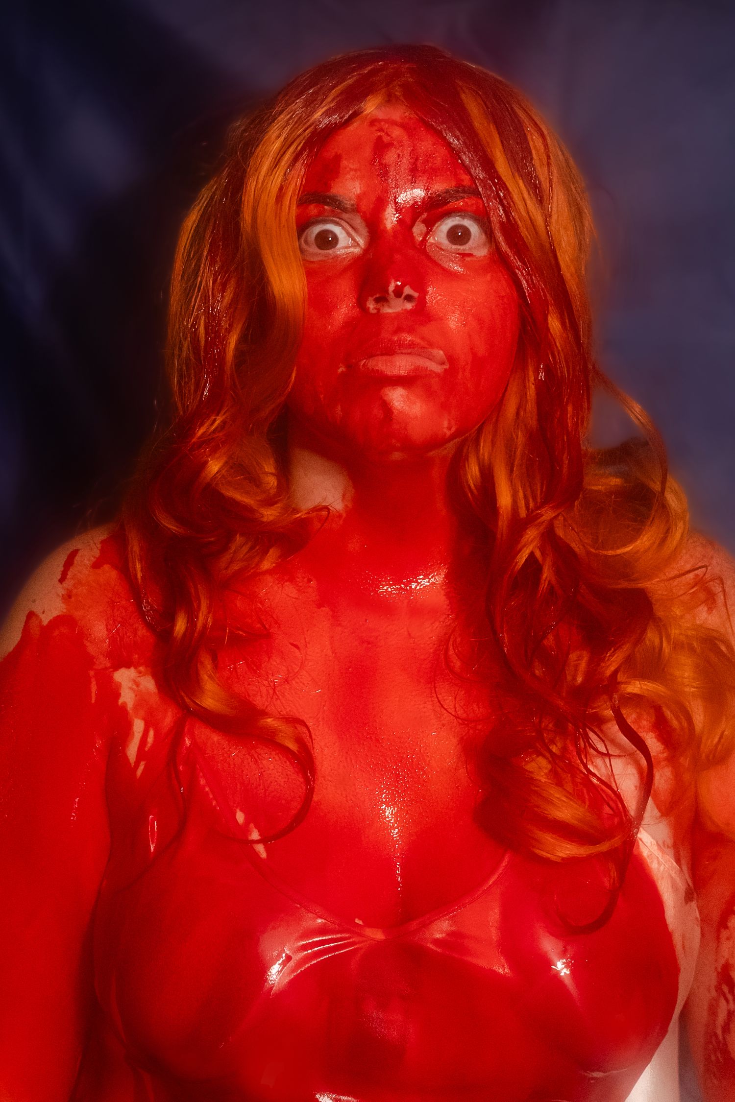 A person covered in fake blood with long wavy hair stares intensely at the camera in dramatic lighting. Carrie inspired halloween photoshoot