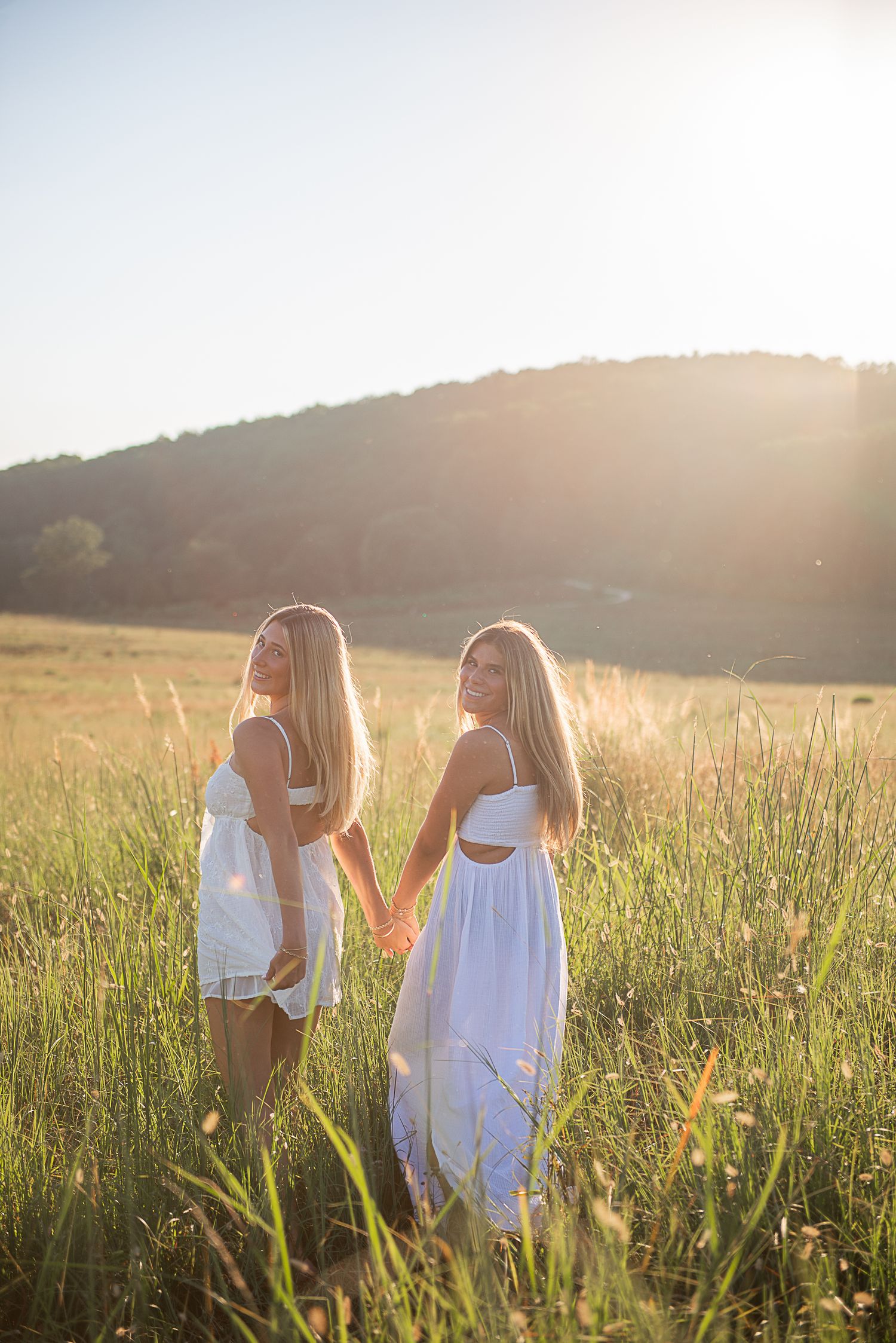 Two people in white dresses dance together in a sunlit meadow at golden hour.