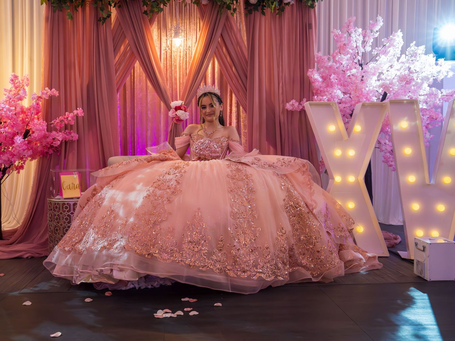 A pink quinceañera dress with intricate lace details and full tulle skirt glows under romantic ballroom lighting.