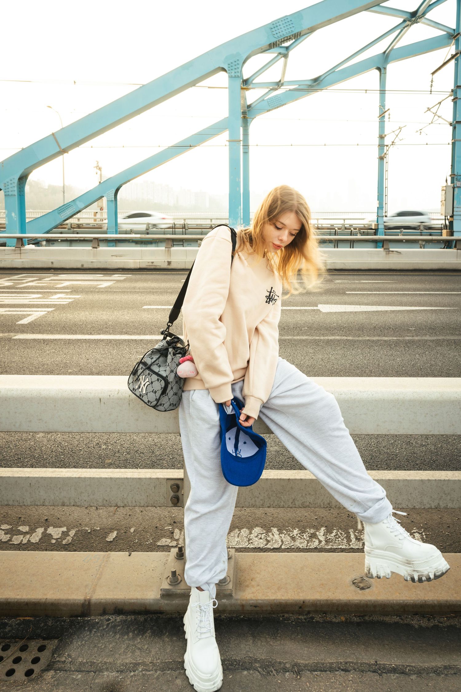 Person in beige jacket and white pants crouches down near a curb while wearing white sneakers.