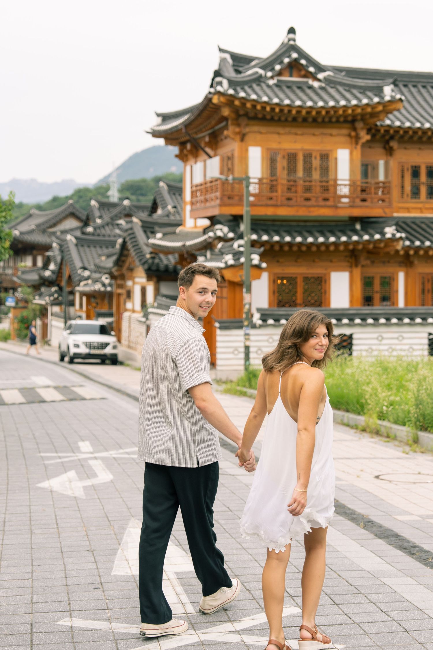 A couple explores traditional Korean hanok village architecture with wooden buildings and stone paths in the background.