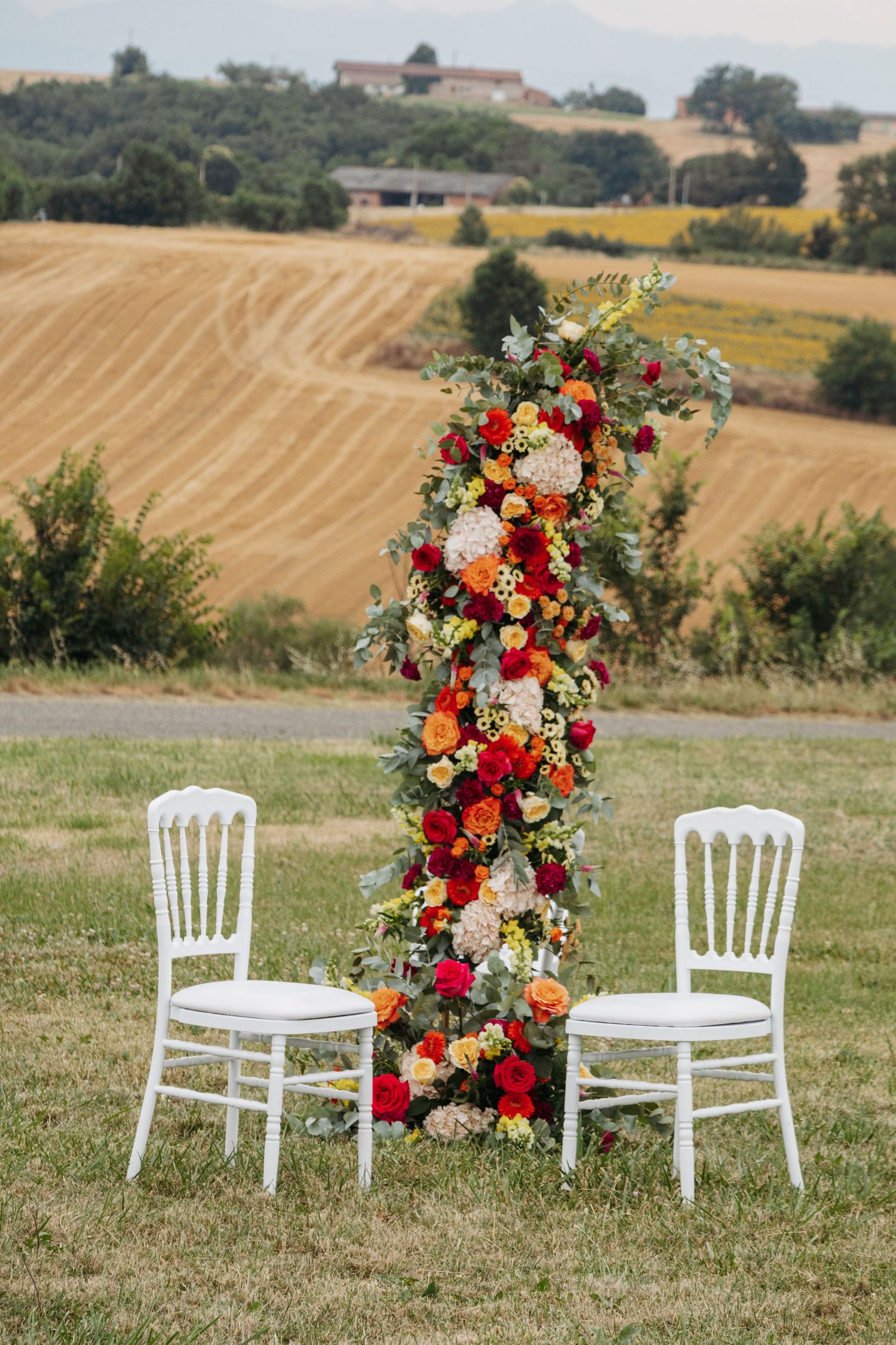 Elegant outdoor wedding ceremony setup with colorful floral arrangement and two white chairs in a sunlit field.