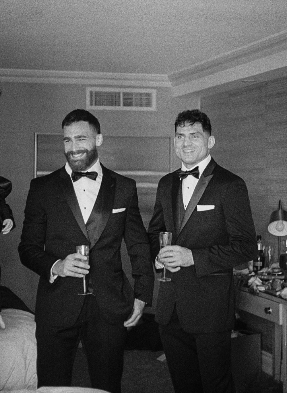 A group in formal black tuxedos and bowties raise champagne glasses in celebration at an elegant indoor event.