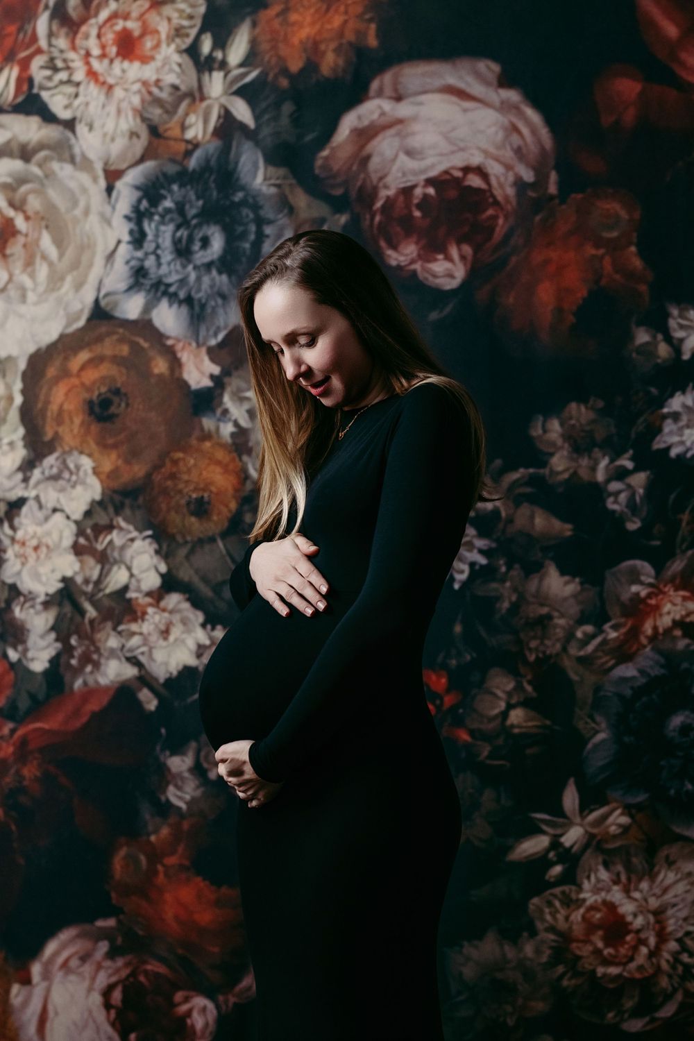 Artistic portrait against dark floral wallpaper showing figure in black clothing with dramatic moody lighting.
