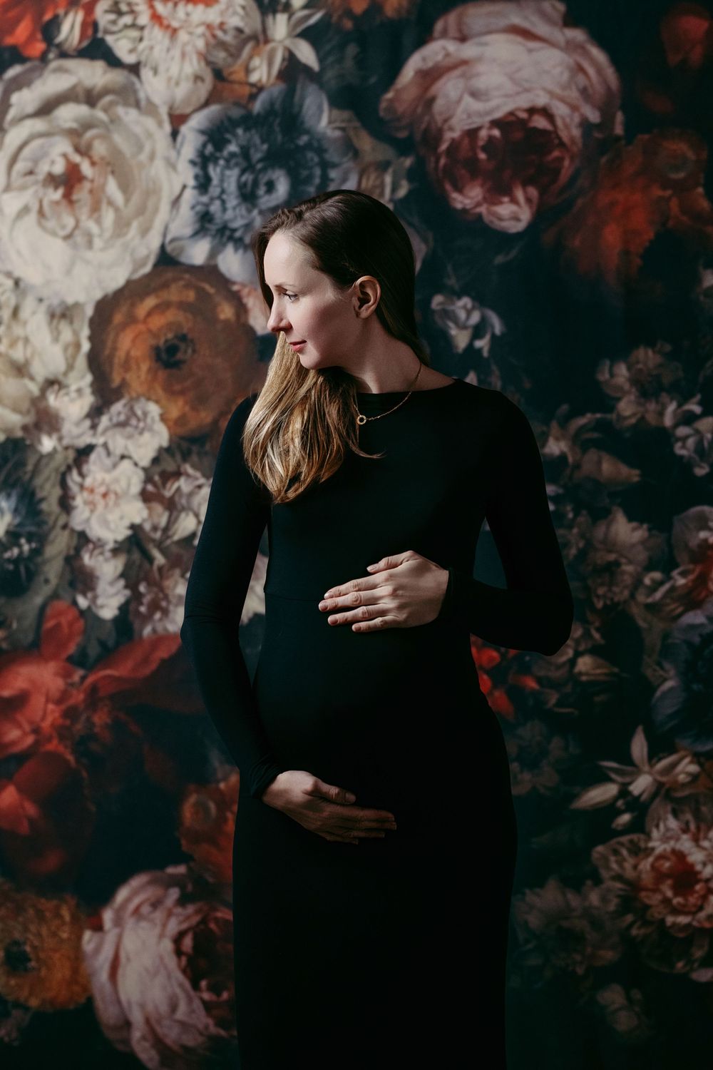 Two people in black clothing embrace intimately against a dark floral wallpaper background.