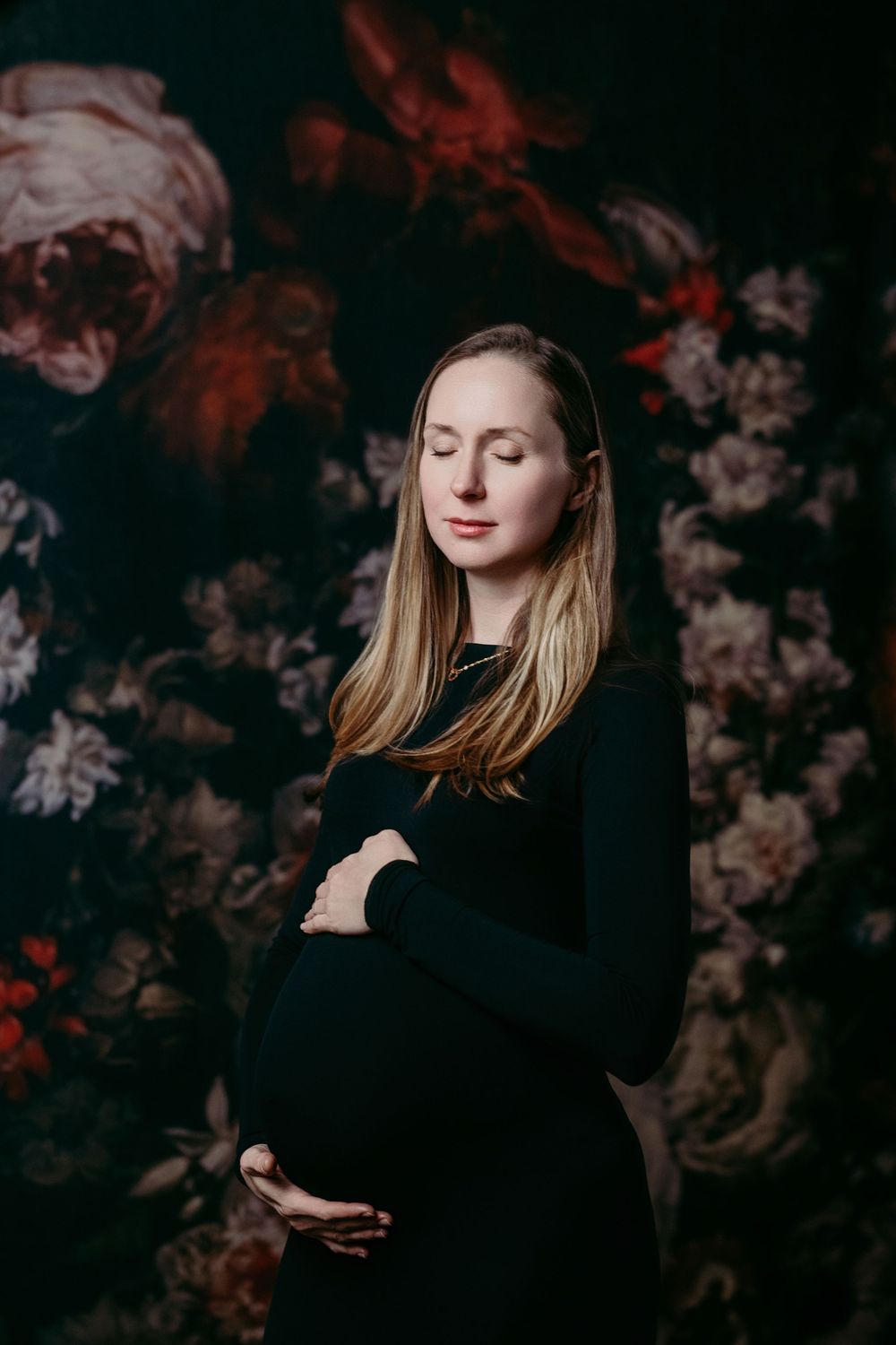 Artistic maternity portrait with dark floral backdrop features a woman in black dress with closed eyes and serene expression.
