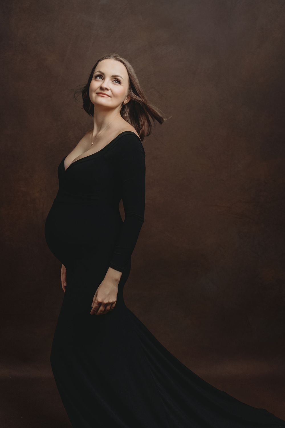 A person in a flowing black dress poses elegantly against a dark brown studio backdrop in a portrait series.